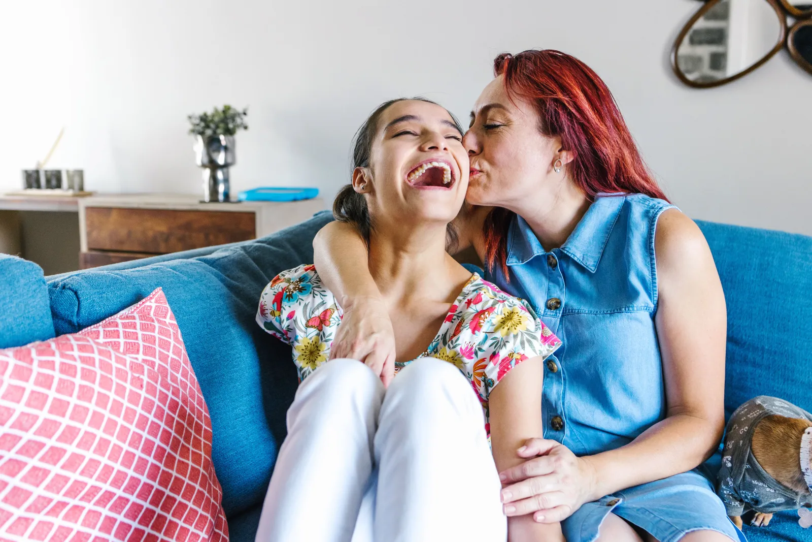 Mother kissing her laughing teenage daughter on the sofa at home
